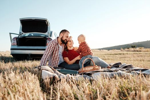 Happy Young Family Mom and Dad with Their Little Son Enjoying Summer Weekend Picnic Sitting on the Plaid Near the Car Outside the City in Field at Sunny Day Sunset, Vacation and Road Trip Concept