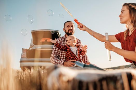 Happy Young Family Mom and Dad with Their Little Son Enjoying Summer Weekend Picnic on Car Outside the City, Playing with Bubbles