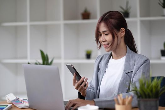 Asian woman designer working using a smartphone blank white computer screen placed at table with color samples