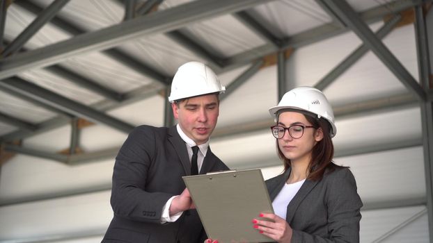 Young man and woman in helmets with documents at a construction site. Chiefs in suits discussing the project of architecture. People look around. 4k