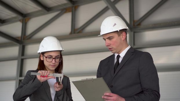 Young man and woman in helmets with documents at a construction site. Businessmen in suits measure the detail with a vernier caliper. 4k