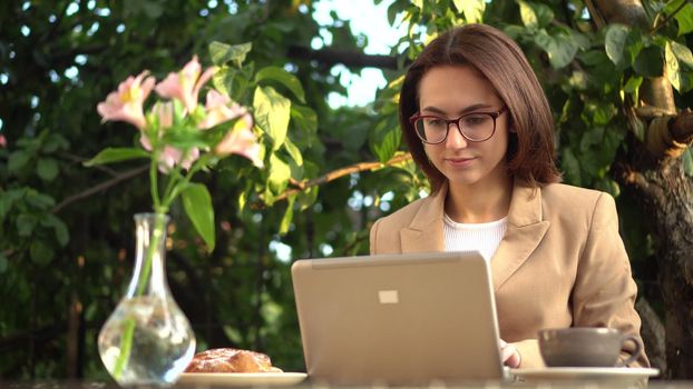 Young business woman sits in a cafe and works at a laptop. Girl with coffee and a bun on the summer veranda. 4k