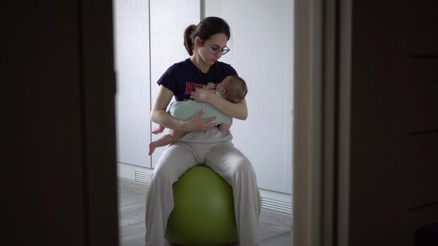 A young mother rocks her baby on a fitness ball. The girl puts the child to sleep at home. 4k