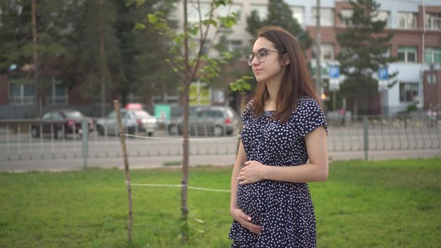 A young pregnant girl walks along the alley and touches her stomach with her hands. Girl with glasses and a dress in the park. 4k