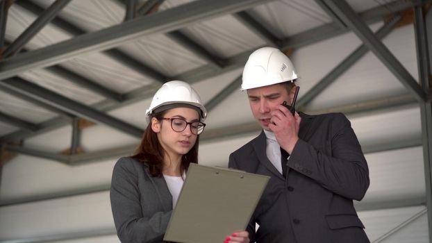 Young man and woman in helmets with documents at a construction site. Bosses in suits are discussing an architecture project. A man speaks on a walkie-talkie. People look around. 4k