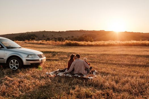 Happy Young Family Mom and Dad with Their Little Son Enjoying Summer Weekend Picnic on the Car Outside the City in Field at Sunny Day Sunset, Vacation and Road Trip Concept