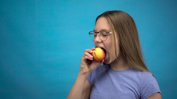Teenager girl with braces on her teeth eats a peach on a blue background. Girl with colored braces bites off a peach. 4k