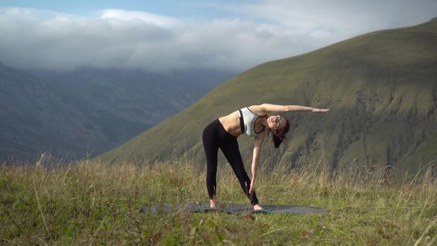 Young woman in tracksuit is engaged in yoga performing triconasana pose in the mountains. The camera moves to create a parallax effect. 4k