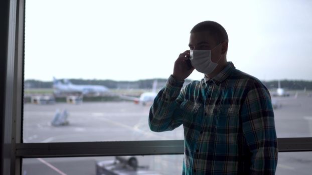 A young man in a medical mask talks on the phone against the background of a window at the airport. Airplanes in the background. 4k