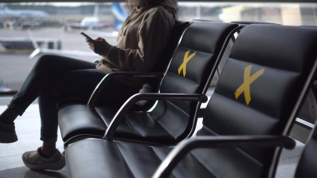 A young woman in a medical mask sits with a phone in her hands against the background of a window at the airport. The chairs are marked with crosses to maintain social distance. 4k