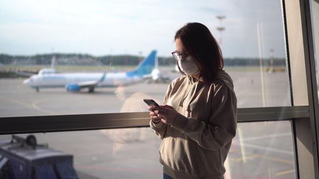 Young woman in a medical mask with a phone in her hands on the background of a window at the airport. Airplanes in the background. 4k
