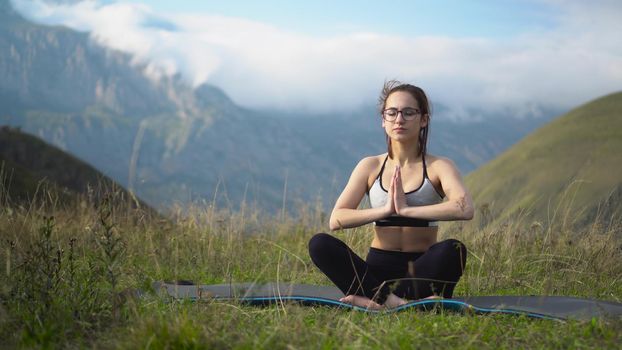 A young woman in a tracksuit practices yoga in the mountains. The camera moves to create a parallax effect. 4k