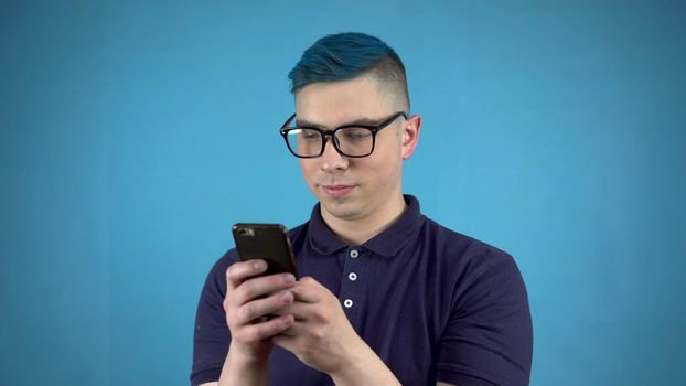 A young man in glasses with blue hair with a phone in his hands. An alternative person is writing in a smartphone on a blue background. 4k