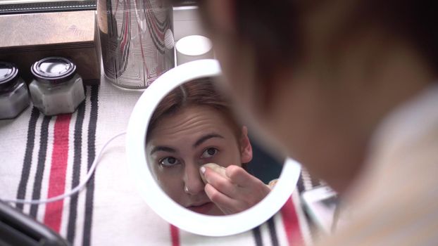 A young woman in front of a window looks into a small mirror with light and applies makeup with a sponge. View of the girl through the mirror. 4k