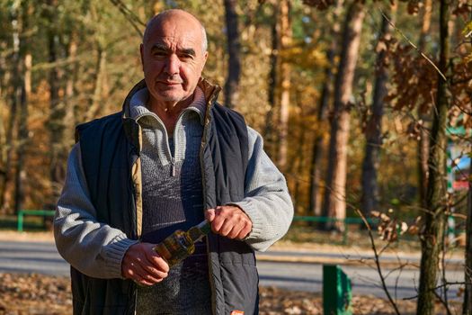 a container, typically made of glass or plastic and with a narrow neck, used for storing drinks or other liquids.. Cheerful elderly male pensioner with a bottle of beer wine alcohol in nature