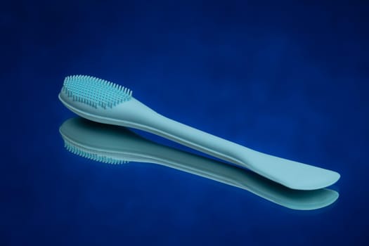 Blue spatula with a brush for applying face mask cream lies on a blue mirror background. High quality photo.
