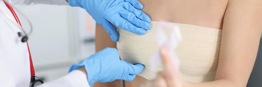 Close-up of doctor examine woman breast and palping it carefully on appointment. Client hold white ribbon sign of breast cancer. Clinic, medicine concept