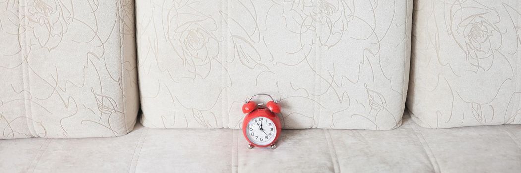 Close-up of beige couch and red vintage clock on it show almost midnight. Decorated christmas tree behind sofa. Get ready to meet new year. Holiday concept