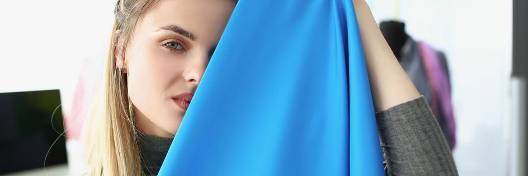 Portrait of young dressmaker woman hiding behind blue high quality fabric. Sewing using premium textile for garment. Atelier, fashion, business concept