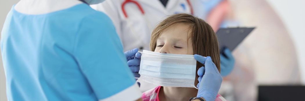 Portrait of medical worker put protective mask on little girl face in clinic. Appointment in clinic with restrictions from covid. Medicine, checkup concept