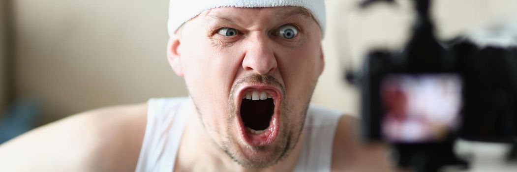 Portrait of man screaming on camera while performing exercise at home. Man in sportswear training indoors, film class on video. Sport, work hard concept