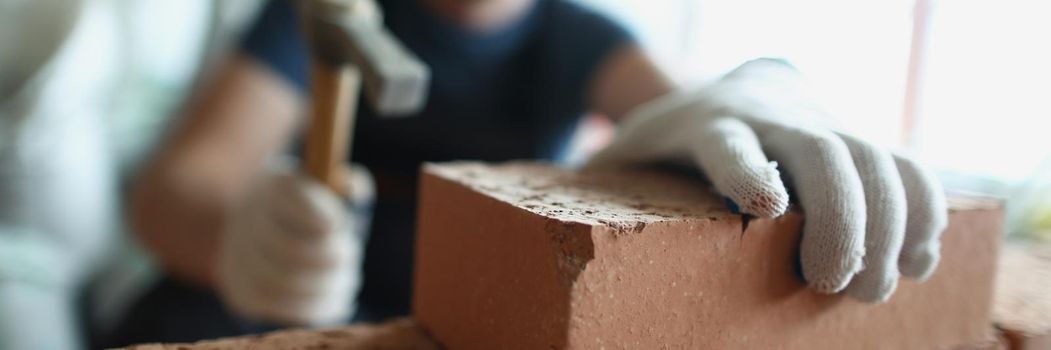 Close-up of skilled foreman in hardhat use hammer equipment to put blocks on unfinished structure. Big concrete wall made of red bricks. Renovation concept