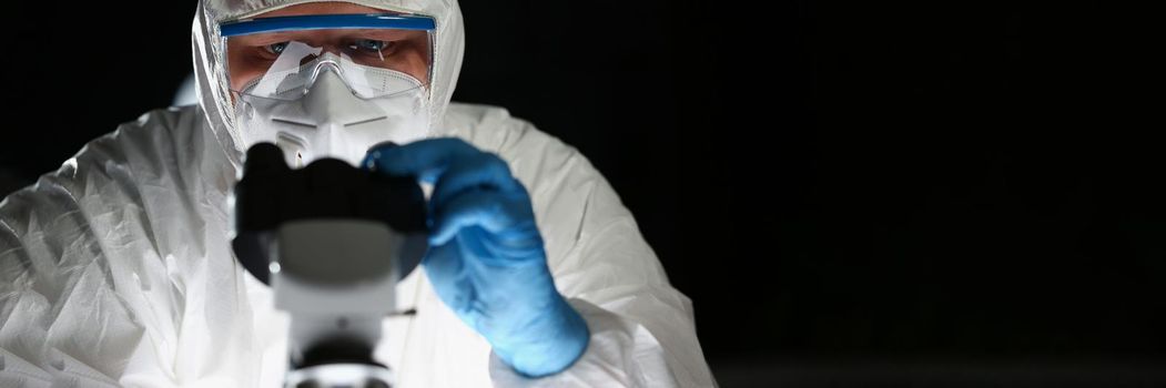 Portrait of male chemist in blue protective gloves look at microscope in chemistry lab. Scientist looking at optical microscopy metal lens. Test concept