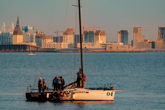 Russia, St. Petersburg, 10 June 2022: The sailing yacht with yachtsmen on board floating to port at sunset, residential complex under construction on the embankment on Vasilievsky island on background. High quality photo
