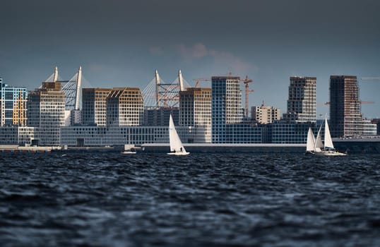 The residential complex under construction on the embankment of the Neva River on Vasilievsky island in sunny day, bulk island, construction cranes, sailing yachts, river navigation. High quality photo
