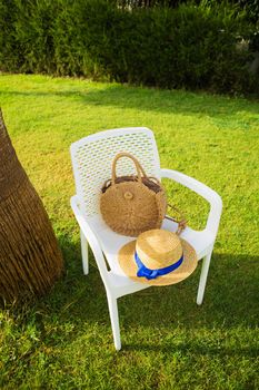 a white chair stands near the palm tree, on the chair a hat and a bag of straw