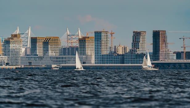 The residential complex under construction on the embankment of the Neva River on Vasilievsky island in sunny day, bulk island, construction cranes, sailing yachts, river navigation. High quality photo