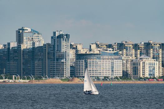 Russia, St. Petersburg, 10 June 2022: The sailing yacht with yachtsmen on board floating at sunset, residential complex under construction on the embankment on Vasilievsky island on background. High quality photo