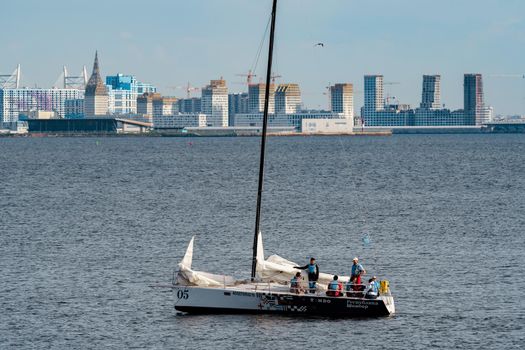 Russia, St. Petersburg, 10 June 2022: The sailing yacht with yachtsmen on board floating at sunset, residential complex under construction on the embankment on Vasilievsky island on background. High quality photo