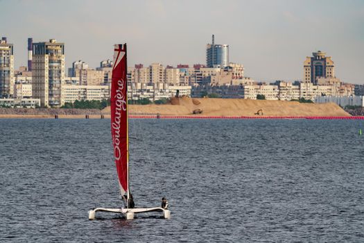 Russia, St. Petersburg, 10 June 2022: The trimaran with professional yachtsmen on board floating at sunset, residential complex under construction on the embankment on Vasilievsky island on background. High quality photo
