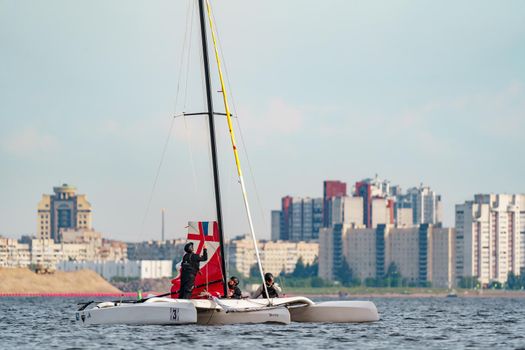 Russia, St. Petersburg, 10 June 2022: The trimaran with professional yachtsmen on board floating at sunset, residential complex under construction on the embankment on Vasilievsky island on background. High quality photo