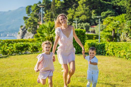 Joyful young mother playing catch together with two little children, boy and girl in green park on a summer day. Childhood, parenting, happiness concept.