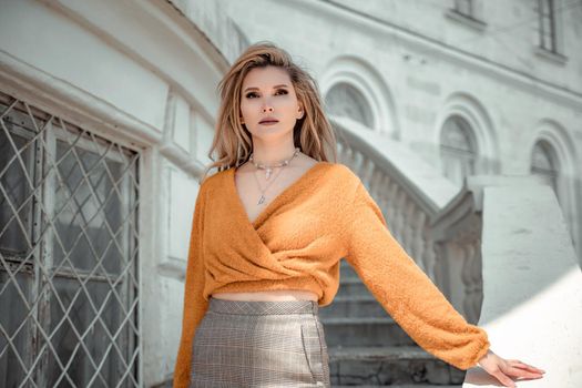 A middle-aged woman looks like a good blonde with curly beautiful hair and makeup on the background of the building. She is wearing a yellow sweater