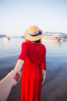 A beautiful girl in a straw hat and red dress is standing on the sea and holding her loved one's hand. Romance, happiness, love.