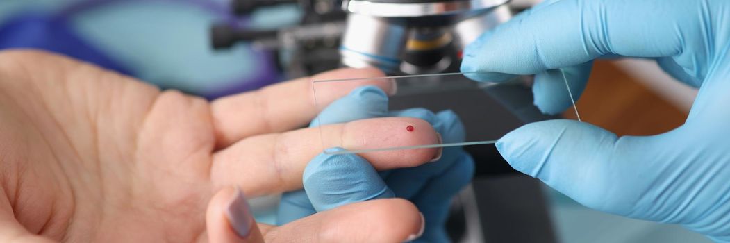 Close-up of laboratory worker taking blood from female finger for investigation. Modern microscope on table in lab. Medicine, chemistry, science concept