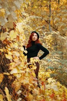 Stylish model among yellow dry foliage of trees. Beautiful female in black beret, knitted sweater and leather skirt. Awesome red-haired woman in autumn park. Walk in fall nature.