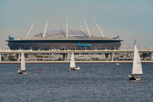 Russia, St. Petersburg, 10 June 2022: Sailing boats return from sailing regatta against the backdrop of the new football stadium of the gas company Gazprom at sunny day. High quality photo