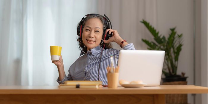 Portrait of Asian Senior Business woman wear headphone and with coffee using video call at morning at home. Account and Finance concept