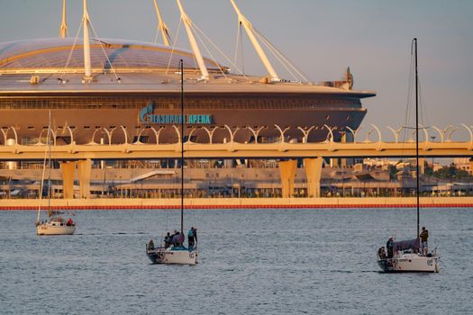 Russia, St. Petersburg, 10 June 2022: Sailing boats return from sailing regatta against the backdrop of the new football stadium of the gas company Gazprom at sunset. High quality photo
