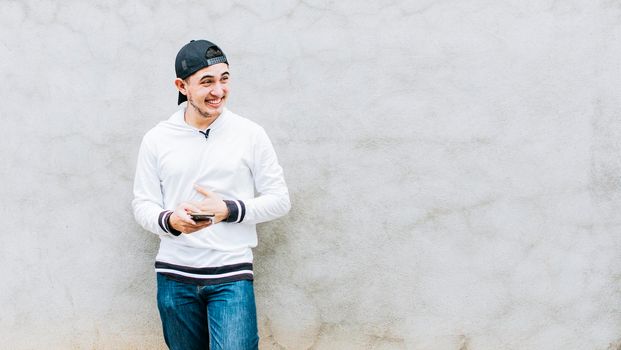 A young latin man laughing with his cell phone leaning against a wall, Handsome guy happy with his cell phone, young latin man happy with his cell phone leaning against a wall