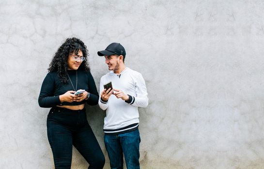 Two smiling friends checking their cell phones, Two teenage friends checking their cell phones and smiling, Two teenagers together checking their cell phones, Guy and girl leaning on a wall checking their cell phones