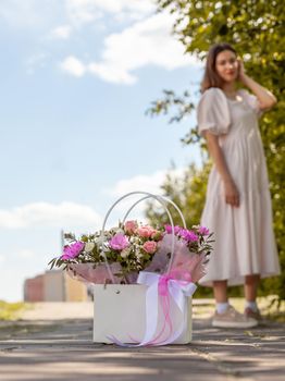 A beautiful bouquet of flowers in a box in the hands of a beautiful girl who walks along the street on a sunny day. Girl in a dress, glasses and sneakers. Focus on the background of flowers.