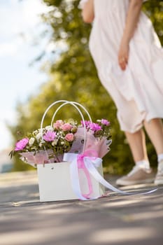 A beautiful bouquet of flowers in a box in the hands of a beautiful girl who walks along the street on a sunny day. Girl in a dress, glasses and sneakers. Focus on the background of flowers.
