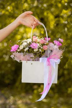 A beautiful bouquet of flowers in a box in the hands of a beautiful girl who walks along the street on a sunny day against the backdrop of green plants. Focus on the background of flowers.