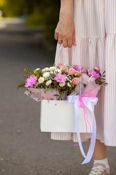 A beautiful bouquet of flowers in a box in the hands of a beautiful girl who walks along the street on a sunny day. Girl in a dress, glasses and sneakers. Focus on the background of flowers.