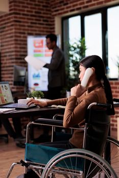 Asian woman with physical impairment talking on landline phone, having remote conversation on office telephone. Wheelchair user with chronic health condition chatting on phone call.